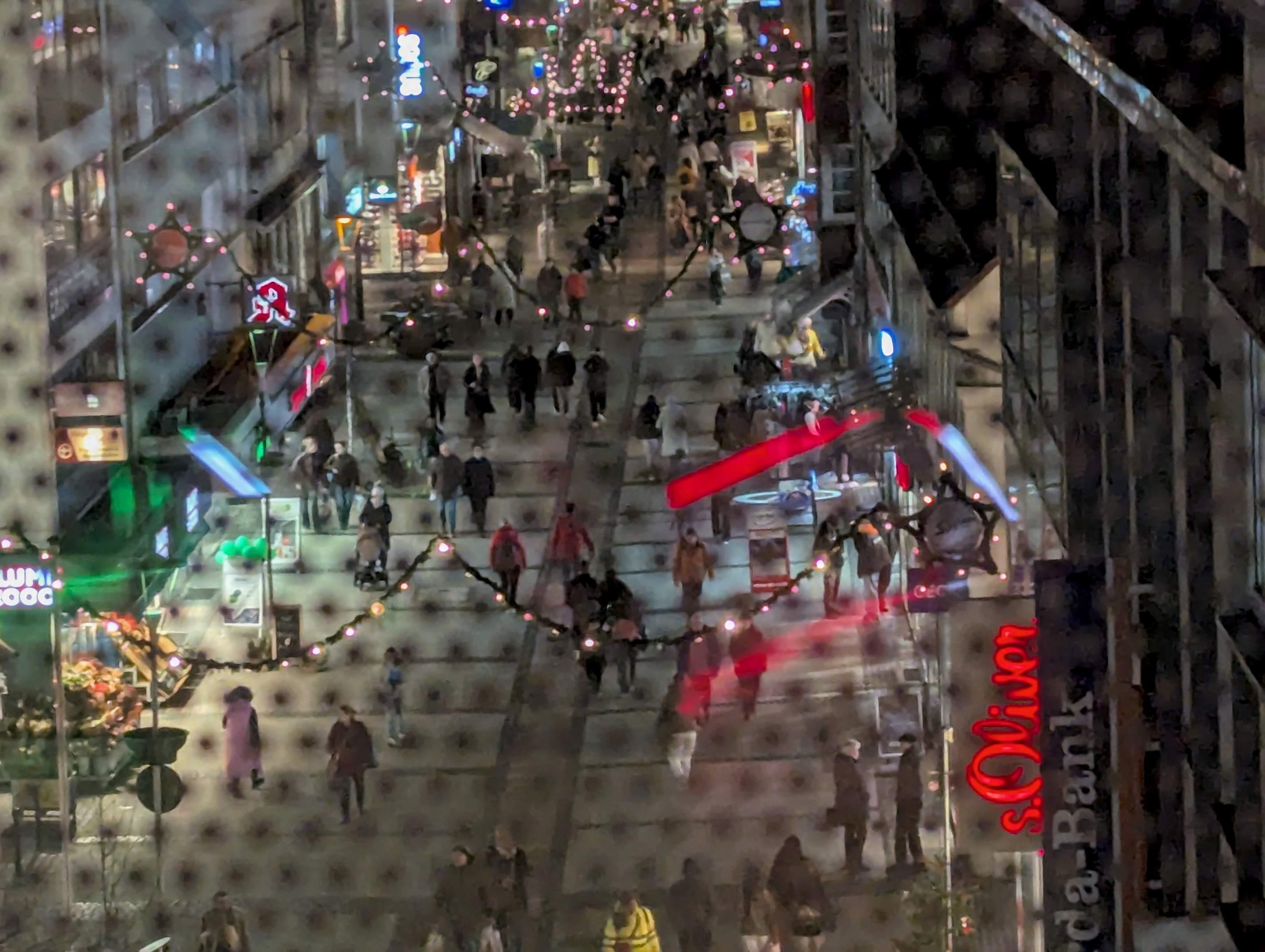 Blick von oben auf eine abendliche, weihnachtlich beleuchtete Einkaufsstraße mit vielen Fußgängerinnen und Fußgängern, bunten Ladenreklamen und Lichterketten, aufgenommen durch ein Fenster mit gepunkteter Struktur im Vordergrund.