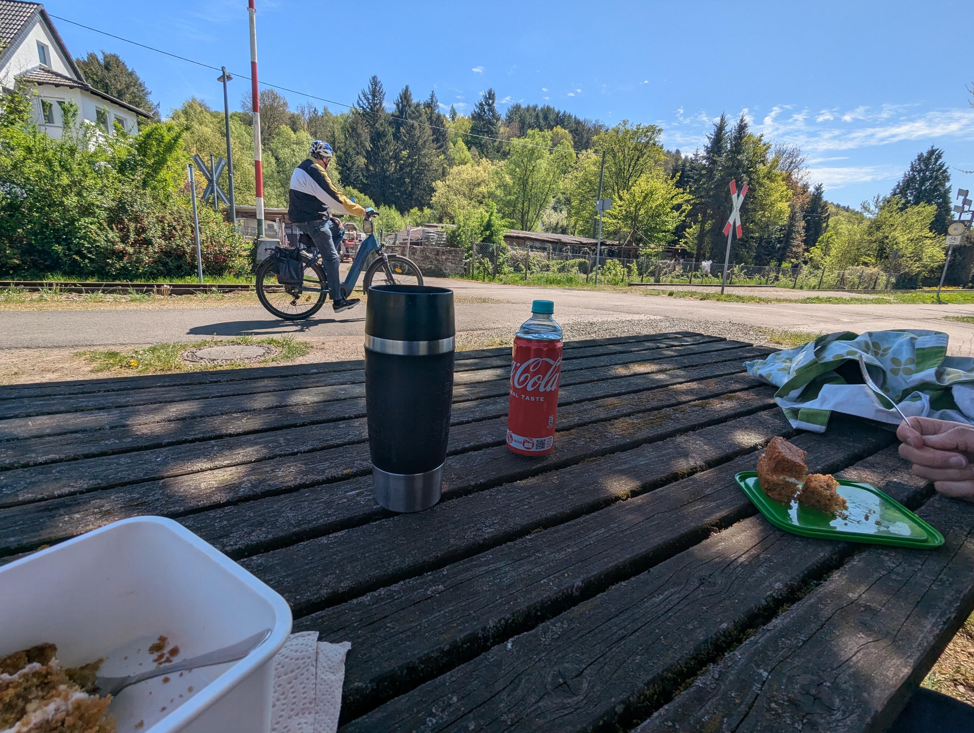 Pause an der Picknickbank an einer Bahnstrecke mit Radfahrer, Getränken und Snack im sonnigen Grünen.
