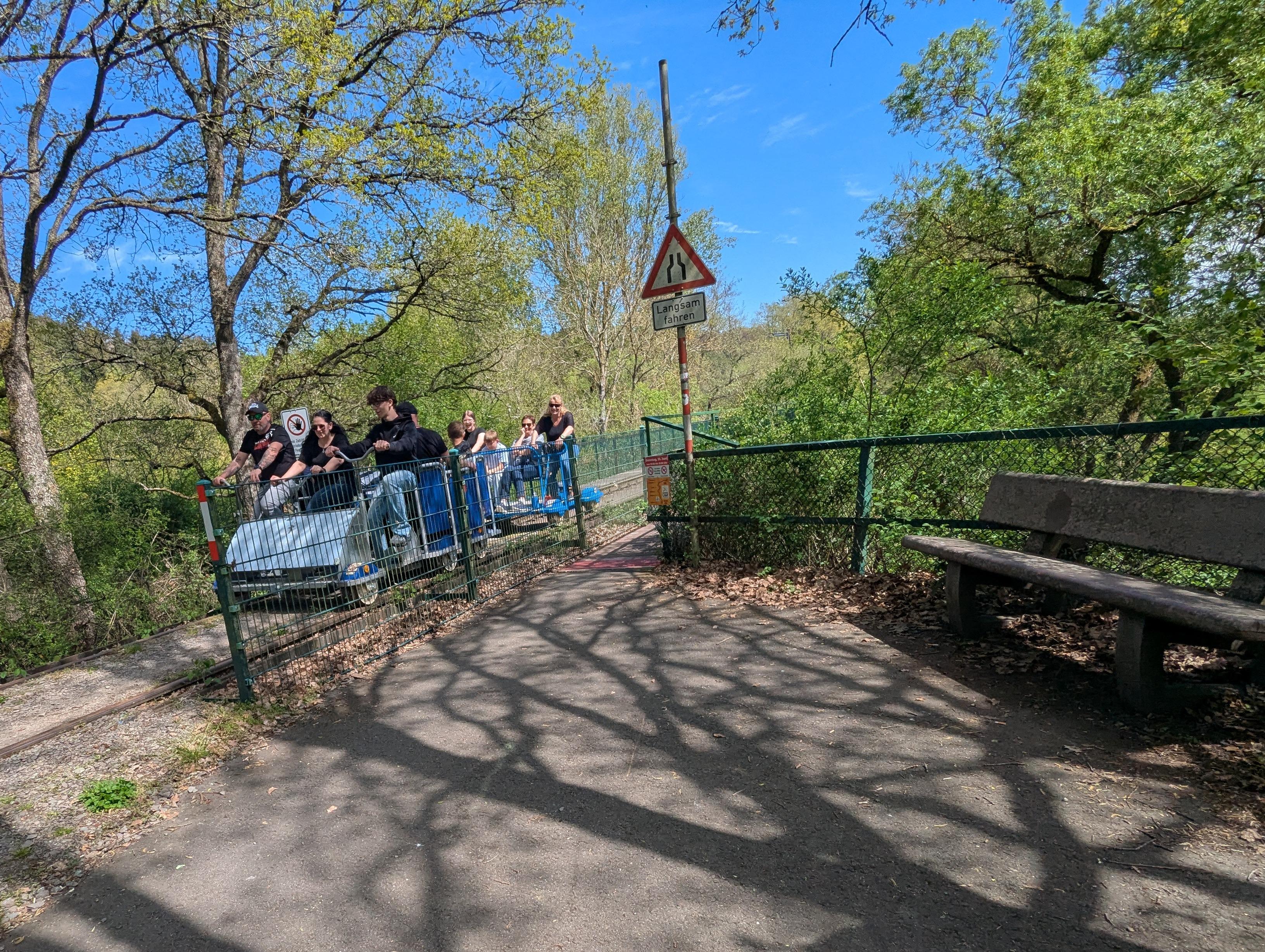 Eine Gruppe von Menschen fährt gemeinsam auf einer Draisine auf einem stillgelegten Bahngleis durch eine bewaldete Umgebung bei sonnigem Wetter. Am Wegesrand steht eine Parkbank und ein Verkehrsschild mit der Aufschrift „Langsam fahren“.